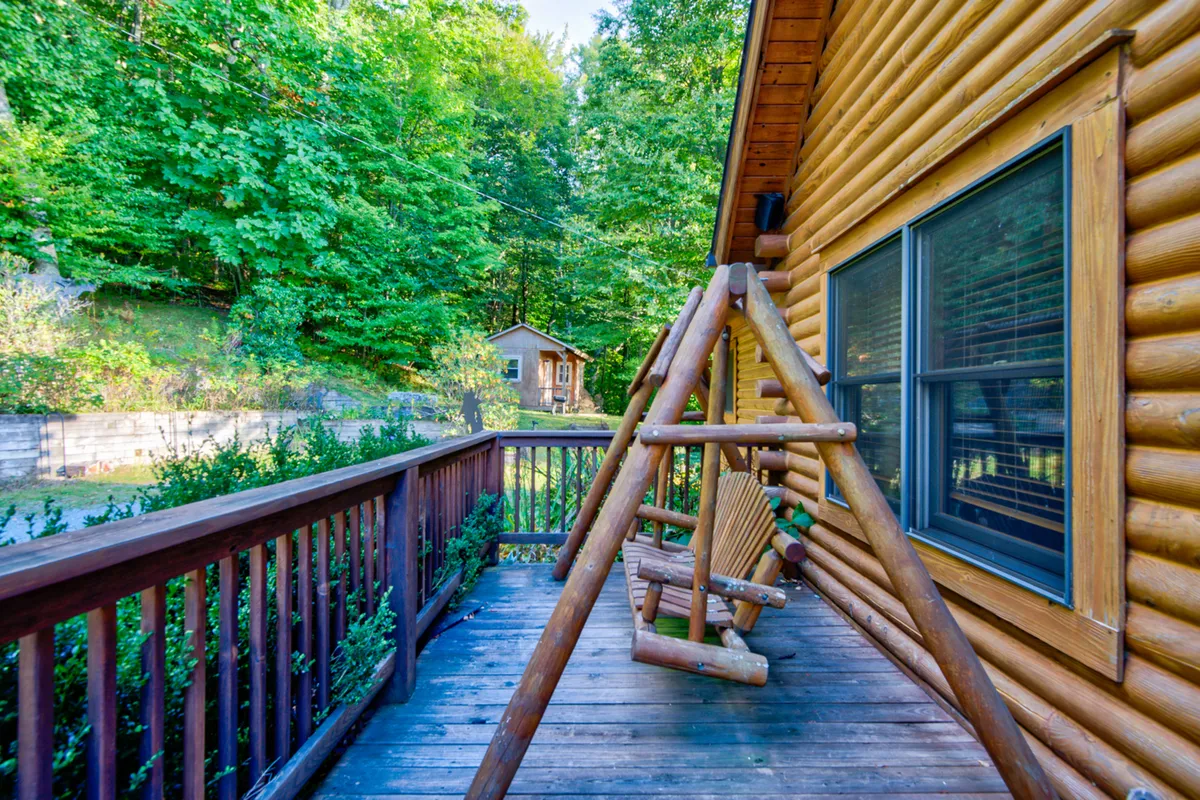 A peaceful outdoor scene with a wooden swing in front of the log cabin, surrounded by green foliage. The inviting swing and tranquil environment suggest a perfect spot for relaxation or watching pets play in the yard.