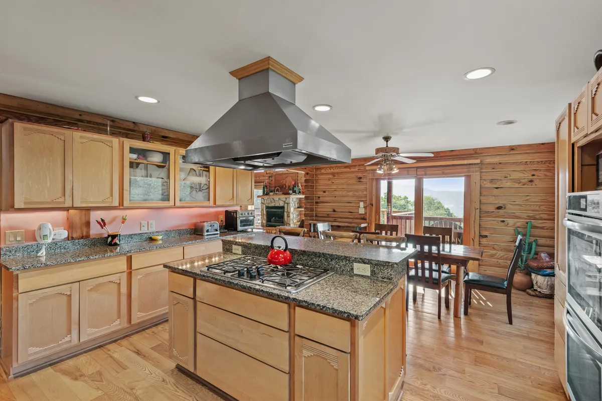 A stylish, well-equipped kitchen featuring a granite island, red kettle, wooden cabinets, and a view toward the dining area, suggesting a space designed for both cooking and socializing with mountain views in the background.