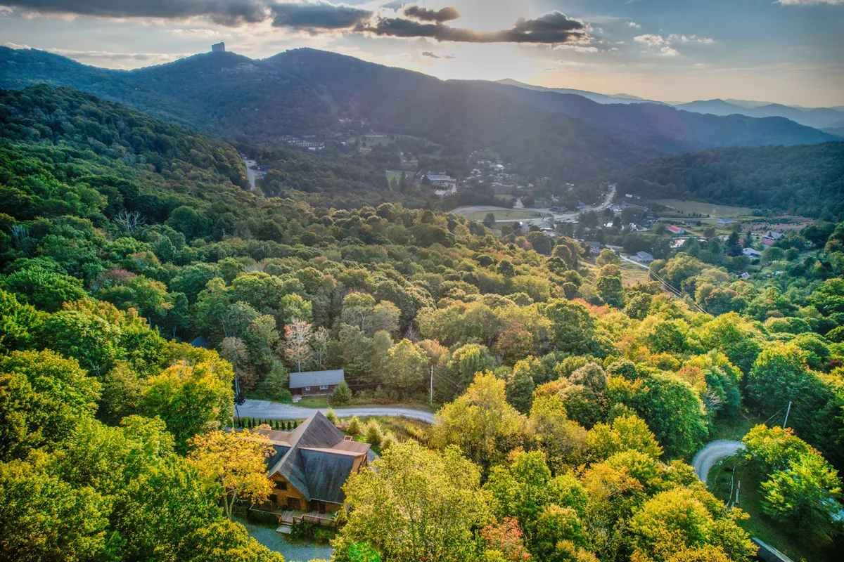 A sweeping view that includes Sugar Bear Lodge, the winding road, forested surroundings, and mountain peaks in the background. The setting sun bathes the landscape in warm light, evoking a sense of adventure and close connection to the natural environment—a reminder that the excitement of Sugar Mountain is just beyond your doorstep.