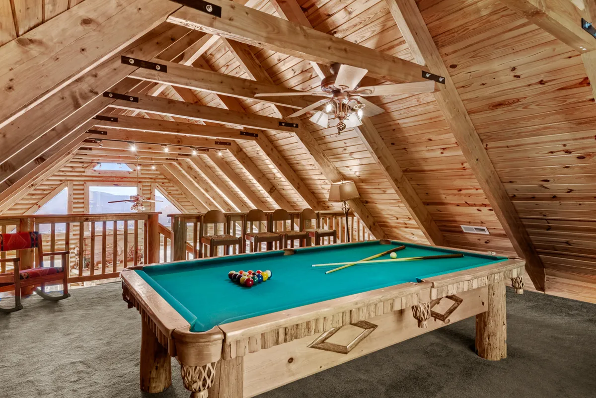 A welcoming interior loft space featuring a pool table as the focal point, surrounded by wooden beams, bar stools, and a ceiling fan. The room has a casual, friendly atmosphere, ideal for playing billiards and socializing with friends or family.