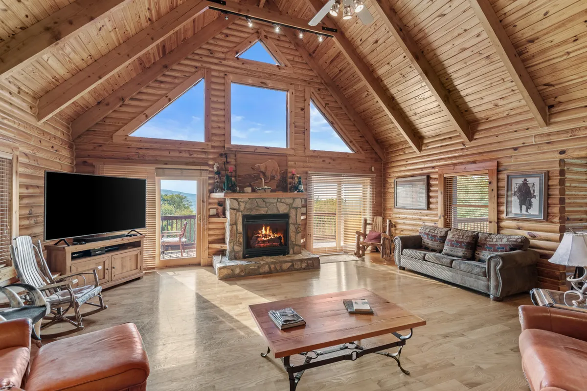 This image displays a spacious lodge living area featuring large windows that frame stunning mountain views, a grand stone fireplace, exposed wooden beams, and plush seating arranged for relaxation. The natural light and scenic views create a strong connection to the outdoors.