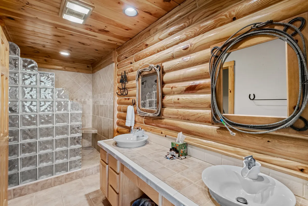 A modern tiled bathroom with rustic log walls, dual sinks, decorative mirrors, and a spacious glass block shower. The room feels both luxurious and functional, ideal for busy mornings or unwinding after a long day.