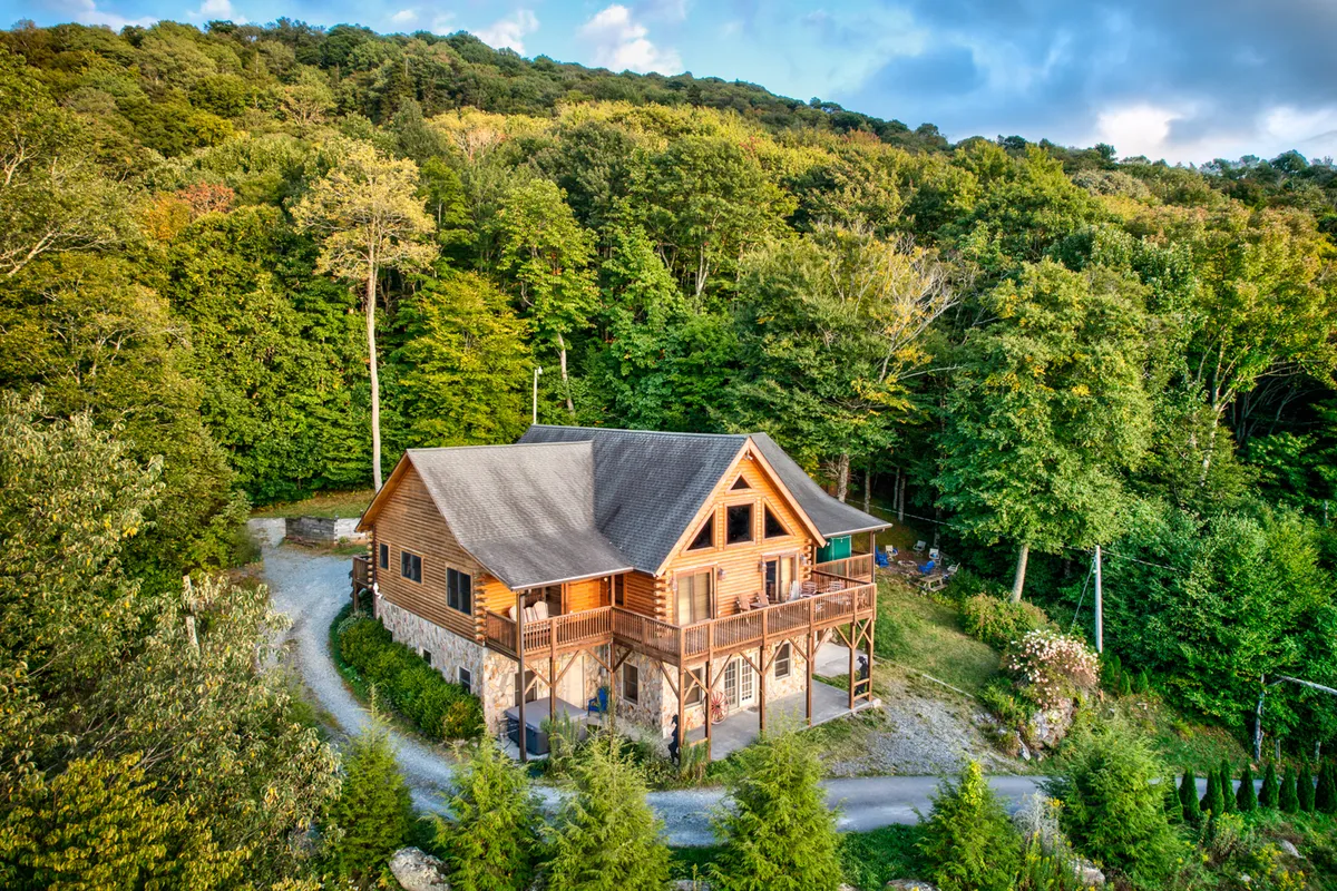 This exterior image showcases the log cabin nestled among the trees with its expansive elevated deck overlooking the tranquil forest. The deck is positioned to catch dramatic sunset views, while the surrounding greenery hints at the endless outdoor adventures available through the seasons.