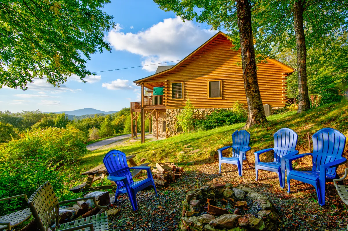 This image features a wooden cabin with a prominent fire pit in the foreground, surrounded by blue chairs and an inviting landscape. The setup clearly encourages social gatherings, storytelling, and evening enjoyment around the warmth of the fire pit.