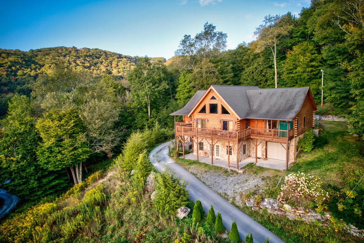 The image shows a majestic log cabin nestled among tall trees, set against a breathtaking mountain backdrop. The cabin's rustic charm is complemented by a spacious driveway and an inviting porch that overlooks sweeping mountain vistas, perfect for relaxing or gathering with loved ones. The soft glow of sunset bathes the scene in warmth, highlighting the natural beauty and tranquil setting of the property.