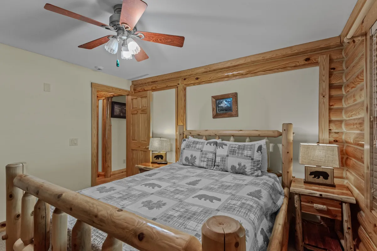 A warmly decorated bedroom with a log bed, bear-themed bedding, wooden bedside tables, and serene natural views just beyond the window. The space feels cozy and welcoming, echoing the rustic charm of the lodge.